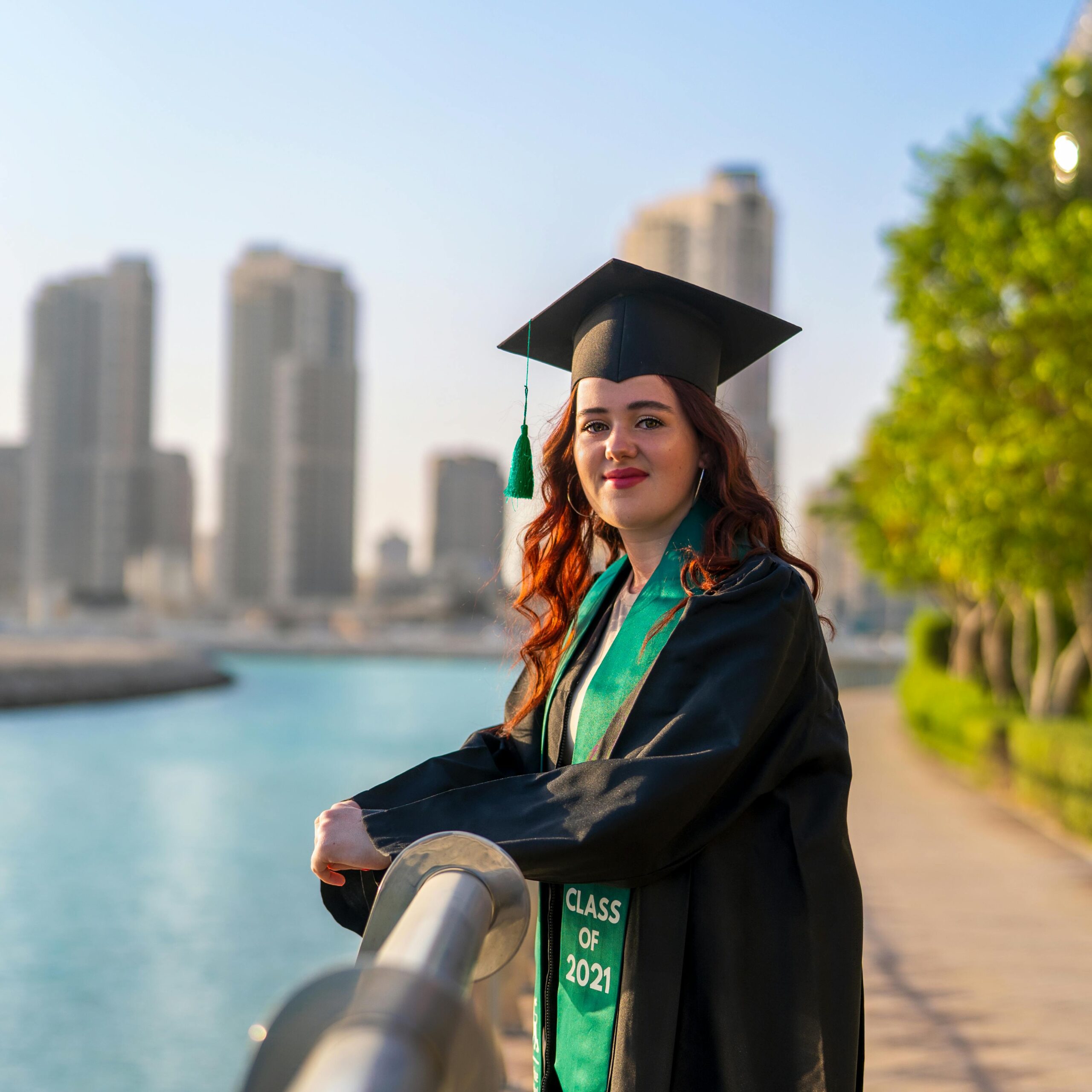 Content female student wearing black graduation cap and gown looking at camera while standing on paved embankment near river in city